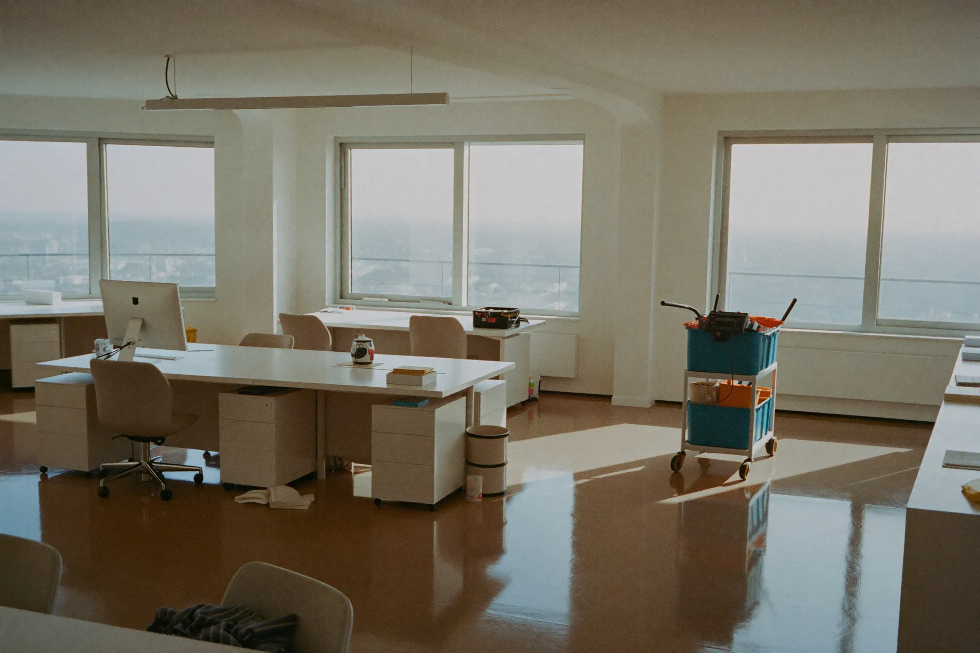 A spotless modern open-plan office with gleaming floors and tidy desks in soft morning light, a cleaning cart parked neatly to one side